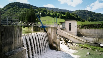 Wasserkraftwerk mit stürzendem Wasser, im Hintergrund Landschaft mit grünen Wiesen und Hügeln voller Bäume
