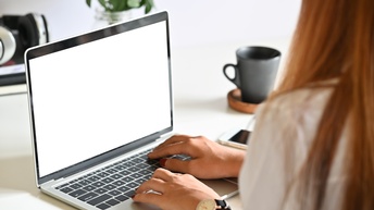 Eine Person tippt auf der Tastatur eines Laptops am Schreibtisch. Daneben stehen eine Dekopflanze, eine Kafeetasse und ein Smartphone.