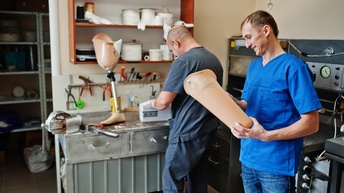 Zwei Personen stehen in Werkstatt: Eine Person blickt lächelnd auf Teil einer Beinprothese, die sie in Händen hält, andere Person in Rückenansicht steht an Tisch auf dem Beinprothese steht, ringsum Werkzeuge, Maschinen und Fertigungsutensilien