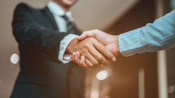 In der linken Bildhälfte ist eine Person in einem Anzug, die einer anderen Person die Hand schüttelt