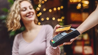 Person mit lockigem blondem Haar und Rosa Shirt bezahlt kontaktlos mit einer Bankomatkarte bei einem Kartenterminal