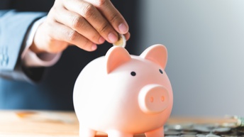 Hand wird Euromünze in ein rosa Sparschwein während weitere Münzen am Tisch verteilt liegen