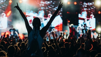 Rückenansicht eines großen Publikums, in dem die Leute teilweise die Hände in die Luft gestreckt haben. Eine Person sitzt auf den Schultern einer anderen Person. Ihnen gegenüber ist eine große Bühne, die beleuchtet wird