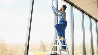 Eine Person steht auf einer Leiter und hält den Stiel eines Gerätes zum Reinigen von Fenstern in den Händen. Er putzt die Fensterscheiben einer Glasfront mit Fenstern, die bis zum Boden reichen. Außen sieht man Nadelbäume. 