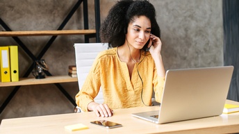 Eine Person mit dunklen, gelockten Haaren sitzt an einem Schreibtisch mit Headset. Vor der Person ist ein aufgeklappter Laptop, auf den sie blickt