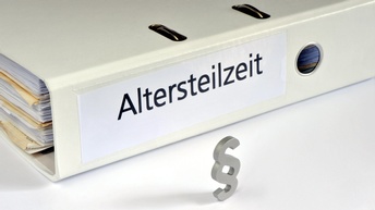 Nahaufnahme eines liegenden, hellen Ordners auf weißem Untergrund. Auf der Seite des Ordners steht in schwarzer Schrift Altersteilzeit. Im Ordner sind Unterlagen. Vor dem Ordner ist ein graues Objekt in Form eines Paragraphenzeichens stehend platziert