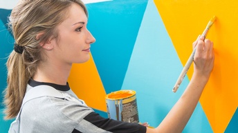 Person steht an Wand und malt gelbblaues Karomuster darauf