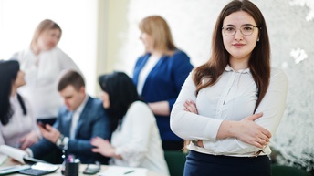 Person im Fokus mit Brillen, in Besprechungszimmer mit vor dem Körper verschränkten Armen stehend, im Hintergrund verschwommen mehrere Personen um Tisch mit Ordnern, Taschenrechnern und anderen Arbeitsutensilien versammelt