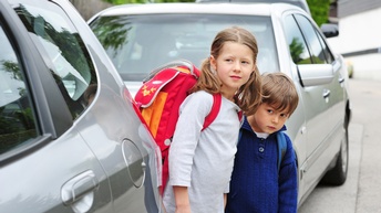 Zwei Kinder mit Schultaschen lugen zwischen zwei Autos am Fahrbahnrand hervor