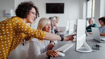 In einem Raum sitzen um einen Tisch sechs Personen. Vor jeder Person ist ein Computer oder ein Laptop. Links vorne im Bild steht eine andere Person neben einer der sitzenden Personen und erklärt etwas am Bildschirm