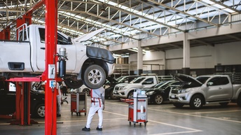 In einer großen Halle parken mehrere Fahrzeuge. Ein Fahrzeug steht erhöht auf einer Hebebühne. Eine Person steht unter dem Fahrzeug