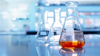 Drei Laborgläser hintereinander aufgestellt, das vorderste Glas enthält braunorange Flüssigkeit