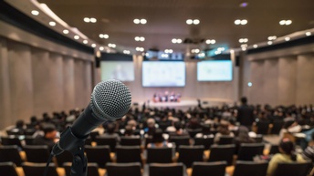 Im Vordergrund ist ein Mikrofon. Im Hintergrund ist ein Saal mit mehreren Sesselreihen, in denen teilweise Leute sitzen. Am Ende des Raums ist eine Bühne, auf der fünf Personen nebeneinander sitzen. Dahinter sind Bildschirme