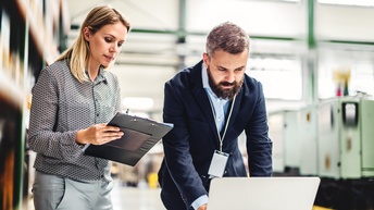 Zwei Personen stehen vor einem niedrigem Tisch in einer Halle. Die linke Person hält ein Clipboard in den Händen und blickt darauf. Die rechte Person beugt sich über den Tisch und hat ihre Hände auf dem vor ihr stehenden, aufgeklappten Laptop