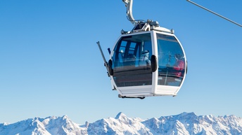 Eine weiße Gondel an Seil hängend, in der Personen in Schikleidung sitzen, an der Gondel außen Schi befestigt, im Hintergrund verschneite Bergkette