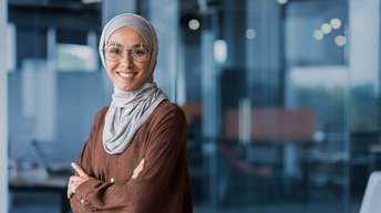Portrait einer lächelnden Person mit kreisrunden Brillen und Kopftuch, im Hintergrund verschwommen Glaswände eines Büros