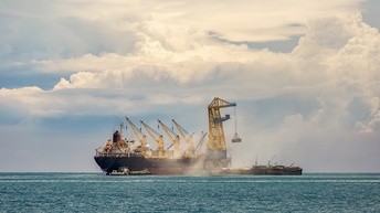Mehrere Frachtschiffe auf dem offenen Meer. Neben ihnen steht ein größeres Schiff, auf dem vier Kräne sind