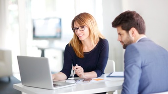 Person mit langen braunen Haaren und Brille sitzt bei einem Schreibtisch mit Unterlagen und blickt auf einen Laptop, eine weitere Person sitzt aufmerksam daneben und blickt ebenfalls auf den Bildschirm