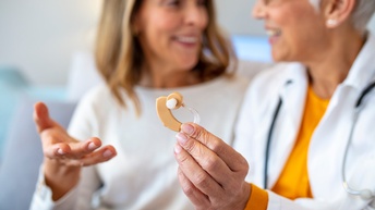 Im Fokus Hand einer Person, die Hörgerät hält, im Hintergrund verschwommen zwei Personen, die einander anblicken im Gespräch