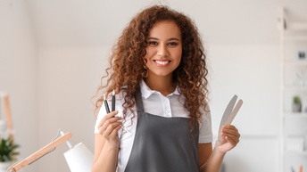 Lächelnde Person mit Locken in grauer Schürze hält verschiedene Feilen in Händen