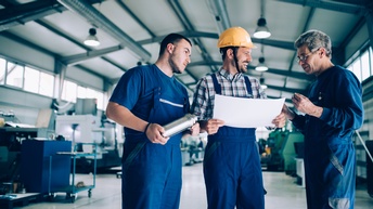 Drei Personen in blauer Arbeitskleidung stehen beisammen und besprechen sich, eine Person trägt einen gelben Schutzhelm und hält ein weißes Blatt Papier, im Hintergrund zeigt sich eine industrielle Werkstätte