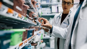 Person mit Brillen in weißem Kittel und mit Stethoskop um den Hals blickt auf Hand weiterer Person in weißem Kittel, die Ampulle hält vor Regal mit Plastikkörben