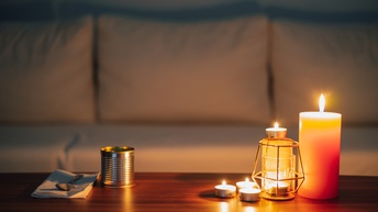 Kerzen und Teelichter sowie eine Konservendose mit Serviette und Löffel liegen auf einem Holztisch und erhellen den dunklen Raum, im Hintergrund steht eine weiße Couch