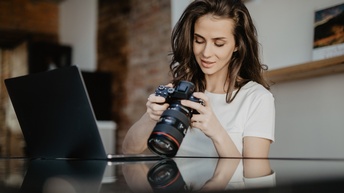Eine Person steht hinter einem hohen Möbelstück, das bis zu ihrer Brust reicht. Die Person hält eine Kamera, auf deren Display sie blickt. Vor ihr ist ein aufgeklappter Laptop.
