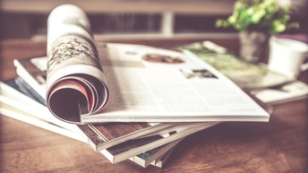 Detailansicht eines Magazinstapels auf Holztisch liegend: oben auf liegendes Magazin offen mit zu Rolle geschlagenen Seiten, im Hintergrund verschwommen Tasse und Vase mit Blumen 