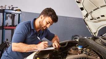 Seitliche Aufnahme einer Person, die sich mit dem Oberkörper leicht nach vorne auf den Rand einer geöffneten Motorhaube lehnt. Unter dem rechten Arm der Person ist ein Klemmbrett, auf dem ein Zettel ist, auf den die Person blickt