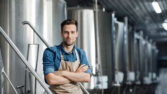 Person mit blauem Hemd und Arbeitsschürze steht stolz mit verschränkten Armen in einer Bierproduktionshalle voller Tanks