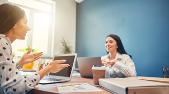Zwei Personen sitzen sich gegenüber auf einem Tisch und unterhalten sich, vor ihnen stehen Laptops sowie liegen Unterlagen auf dem Tisch, im Hintergrund ist eine blaue Wand