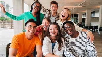 Drei junge Personen sitzen an einem Tisch an einer überdachten Stelle im Freien. Hinter ihnen sind fünf weitere Personen, die sich über die sitzenden Personen lehnen. Jede Person hat den Mund geöffnet und die Mundwinkel nach oben gezogen