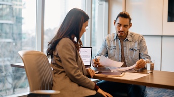 Zwei Personen sitzen sich an einem Tisch gegenüber. Vor der linken Person sind ein Laptop und Tablet. Die andere Person hält Zettel. Beide blicken auf die Zettel.
