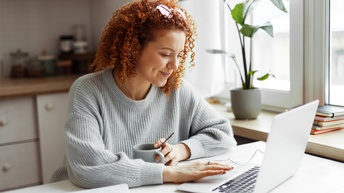 Person sitzt an Tisch in Wohnküche und blickt auf aufgeklappten Laptop vor sich