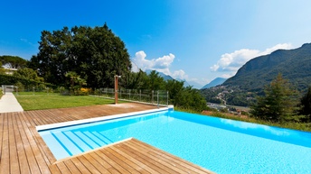 Ein großes Becken mit klarem Wasser, in das Stiegen an einer Stelle führen, wird von Holzbrettern umgeben. Links von den Brettern ist eine grüne Rasenfläche. Im Hintergrund sind grüne Berge und Häuser