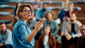 Leicht seitliche Aufnahme einer Person, die sich nach rechts aufdreht und spricht. In der linken Hand hält sie ein Mikrofon. Mit der rechten Hand deutet sie. Der Person gegenüber sitzen mehrere Personen in einem Hörsaal