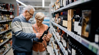 Zwei Personen mittleren Alters stehen in einem Gang eines Supermarktes und betrachten eine Flasche Wein