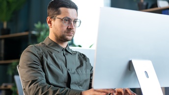 Eine Person mit kurzen Haaren, Bart und Brille sitzt an einem Tisch und tippt auf einer Tastatur eines Computers