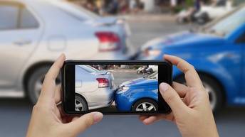 Zwei Hände einer Person halten horizontal ein Smarthone, auf dessen Bildschirm zwei hintereinander stehende Autos sind. Das hintere Auto berührt das vordere Auto. Im Hintergrund sind die beiden Fotos der Aufnahme