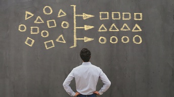 Eine Person ist von hinten fotografiert und steht vor einer schwarzen Tafel. Darauf sind mit gelber Kreide links Rechtecke, Dreiecke und Kreise gezeichnet. In der Mitte ist ein Strich, von dem drei Pfeile ausgehen, die Symbole sind rechts in einer Reihe