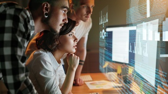 Seitliche Aufnahme einer an einem Schreibtisch sitzenden Person. Daneben stehen zwei weitere Personen, die sich am Tisch abstützen. Über dem Schreibtisch schwebt eine Projektion eines Bildschirms, auf dem Quellcodes und Statistiken sind