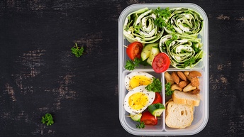 Top-Shot einer Plastikbox in der rechten Hälfte mit drei Fächern. In jedem Fach ist unterschiedliches Essen, zum Beispiel Gurken, Tomaten, Nüsse, Zwieback, Wraps mit Gemüse