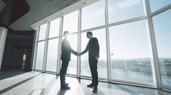 Zwei Personen in Anzügen stehen  vor einer Glasfront und schütteln sich die Hände. Durch die Glasscheiben scheint die tiefstehende Sonne