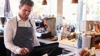 Sanft lächelnde Person in Schürze blickt und tippt auf Display in Barbereich, im Hintergrund Kaffeemaschine, Teller und Tassen