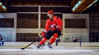 Eine Person in Eishockey-Ausrüstung fährt mit Schlittschuhen auf einer Eisfläche in einer Halle und manövriert einen Puck mit einem Schläger.