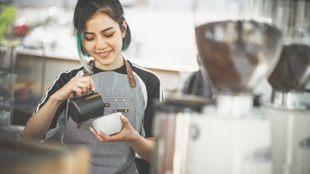 Frontale Aufnahme einer lächelnden Person in Schürze, die hinter einem Tresen steht. Die Person hält mit der linken Hand eine weiße Tasse. Über die Tasse hält sie eine Milchkanne.