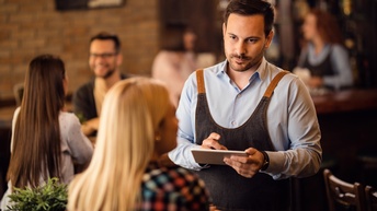 Rückenansicht einer Person, die an einem Tisch in einem Lokal sitzt. Ihr gegenüber steht eine Person mit Schürze, kurzen Haaren und Bart, die ein Tablet hält und zu der sitzenden Person blickt