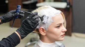 Seitliche Aufnahme einer sitzenden Person mit blonden Haaren und Umhang, die geradeaus blickt. Eine andere Person trägt mit einem Pinsel Färbemittel auf dem Kopf auf