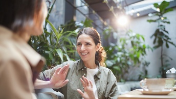 Zwei Personen sitzen sich gegenüber. Die zur Kamera gewandte Person gestikuliert mit den Händen. Vor ihr ist ein Tisch, auf dem Tassen stehen. Im Hintergrund sind grüne Pflanzen und eine Fensterfront.